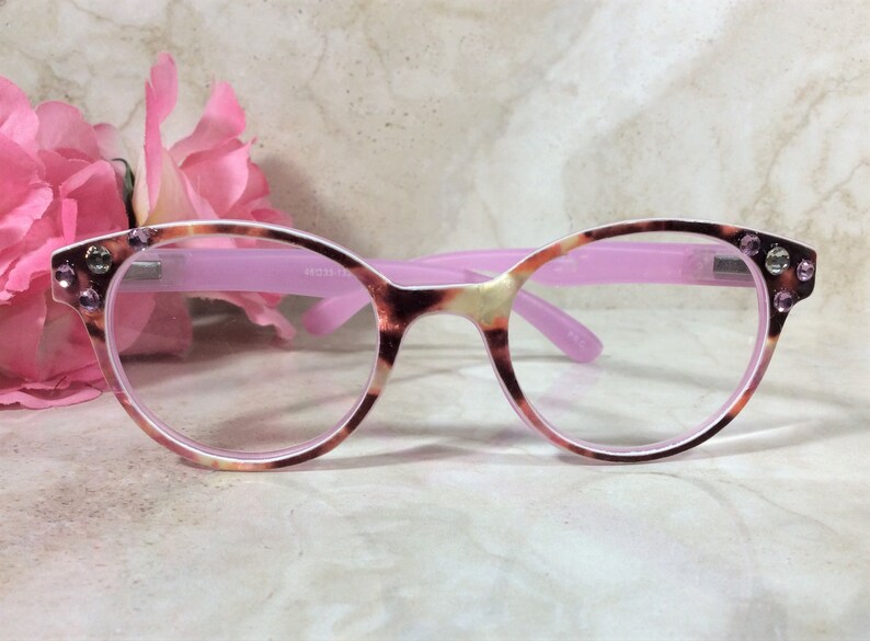 May include: A pair of eyeglasses with a pink frame and a tortoiseshell pattern on the rims. The temples are also pink. The corners of the frames are embellished with small, sparkling rhinestones. The lenses are clear.