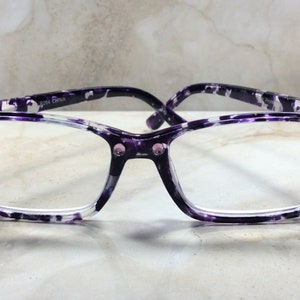 K&ouml;nnte beinhalten: Brille mit rechteckigem Rahmen und lila-wei&szlig;em Muster. Die Brille hat kleine rosa Strasssteine an den Ecken und am Steg. Die B&uuml;gel sind dunkelviolett.