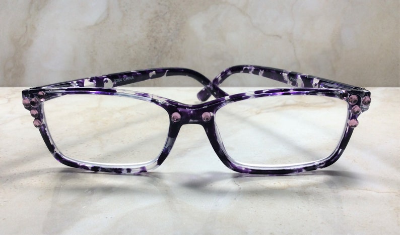 K&ouml;nnte beinhalten: Brille mit rechteckigem Rahmen und lila-wei&szlig;em Muster. Die Brille hat kleine rosa Strasssteine an den Ecken und am Steg. Die B&uuml;gel sind dunkelviolett.