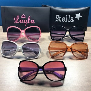 May include: An assortment of sunglasses in various colors, including pink, black, white, and orange, with gradient lenses. Two black cases are visible, one with the name "Layla" and a crown, and the other with the name "Stella" and a star.