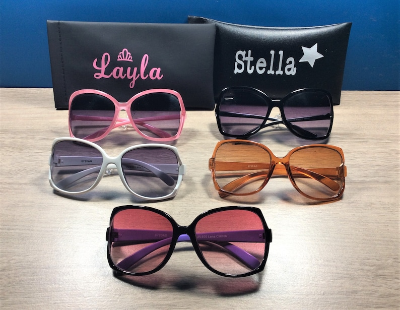 May include: An assortment of sunglasses in various colors, including pink, black, white, and orange, with gradient lenses. Two black cases are visible, one with the name "Layla" and a crown, and the other with the name "Stella" and a star.