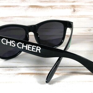 May include: Black sunglasses with dark lenses. The text "CHS CHEER" is printed in white on the left arm of the sunglasses. The sunglasses are resting on a light-colored wooden surface.
