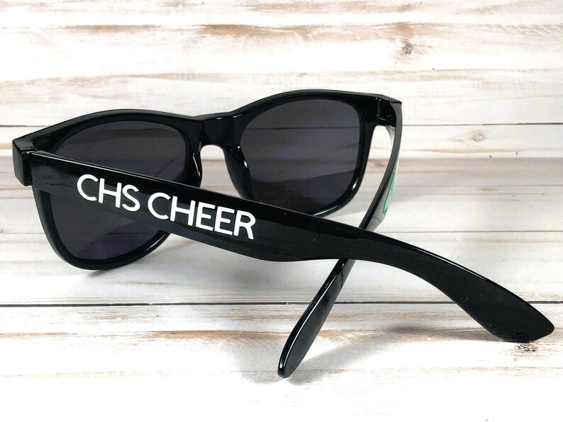 May include: Black sunglasses with dark lenses. The text "CHS CHEER" is printed in white on the left arm of the sunglasses. The sunglasses are resting on a light-colored wooden surface.