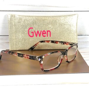 Pu&ograve; includere: Occhiali con motivo floreale rosso, nero e bianco, con lenti trasparenti e decorazioni con strass. Sono appoggiati su una superficie dorata, con una custodia dorata con paillettes dietro con il nome "Gwen" in rosa.