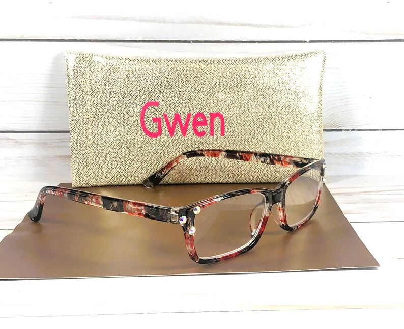 Pu&ograve; includere: Occhiali con motivo floreale rosso, nero e bianco, con lenti trasparenti e decorazioni con strass. Sono appoggiati su una superficie dorata, con una custodia dorata con paillettes dietro con il nome "Gwen" in rosa.