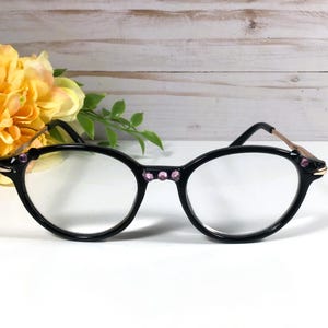 May include: Black oval eyeglasses with clear lenses. The frames have pink jewel accents and gold-coloured temples. A bouquet of yellow flowers is in the background, with a white and light brown wooden surface.
