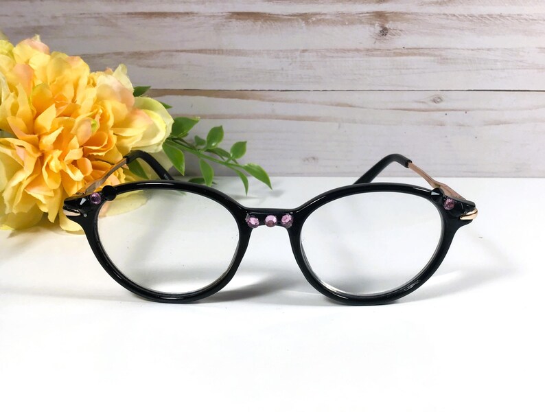May include: Black oval eyeglasses with clear lenses. The frames have pink jewel accents and gold-coloured temples. A bouquet of yellow flowers is in the background, with a white and light brown wooden surface.