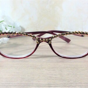 May include: Maroon-framed spectacles with a cat-eye design, embellished with gold-toned beads along the upper rim. The lenses are clear, and the glasses are set against a soft, neutral background.
