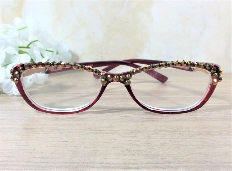 May include: Maroon-framed spectacles with a cat-eye design, embellished with gold-toned beads along the upper rim. The lenses are clear, and the glasses are set against a soft, neutral background.