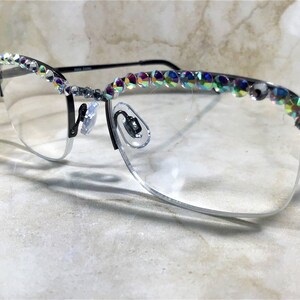May include: Half-rimmed eyeglasses with a black frame and clear lenses. The top of the frame is embellished with iridescent rhinestones. The glasses have a silver-colored nose bridge and temples. The image is taken at an angle, showing the glasses resting on a light-colored surface.