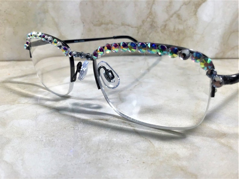 May include: Half-rimmed eyeglasses with a black frame and clear lenses. The top of the frame is embellished with iridescent rhinestones. The glasses have a silver-colored nose bridge and temples. The image is taken at an angle, showing the glasses resting on a light-colored surface.