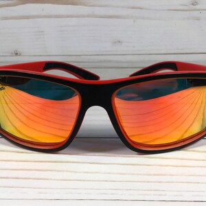 K&ouml;nnte beinhalten: Schwarze und rote Sonnenbrille mit reflektierendem orangefarbenem Glas. Die Sonnenbrille hat ein sportliches Design.
