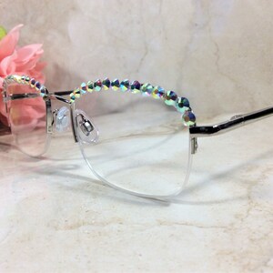 May include: Silver-toned semi-rimless eyeglasses featuring a row of iridescent, multi-colored crystals along the top of the frames. The clear lenses are held by a thin metal structure with adjustable nose pads and slender temples extending outward.