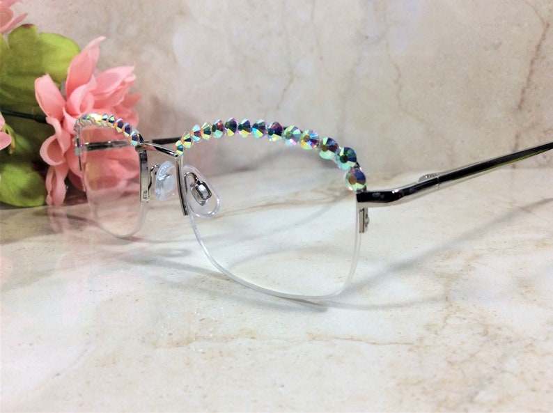 May include: Silver-toned semi-rimless eyeglasses featuring a row of iridescent, multi-colored crystals along the top of the frames. The clear lenses are held by a thin metal structure with adjustable nose pads and slender temples extending outward.