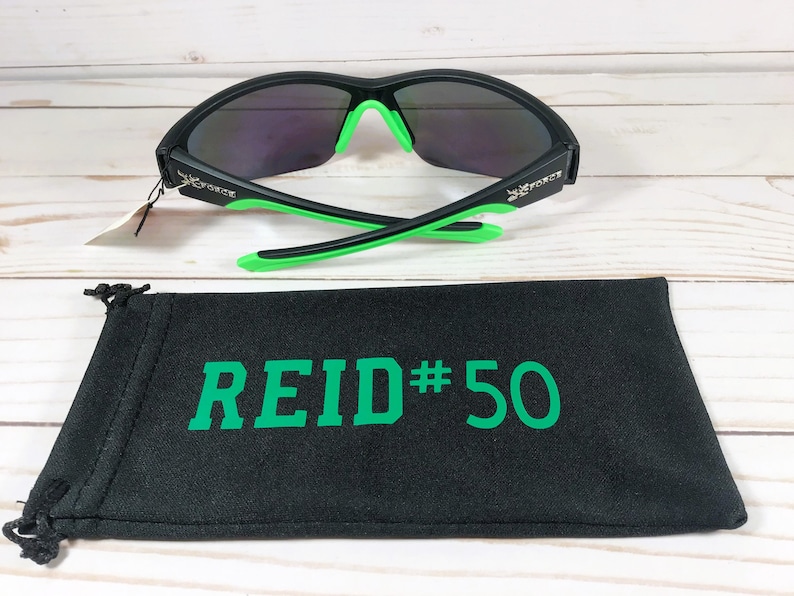 May include: Black sunglasses with green accents and mirrored lenses. The sunglasses are paired with a black drawstring bag with the text "REID#50" in green. The sunglasses have the word "FORCE" on the side.