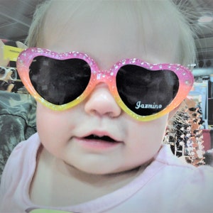 May include: A young girl wearing pink and yellow heart-shaped sunglasses with the word "Jazmino" printed on the lenses.