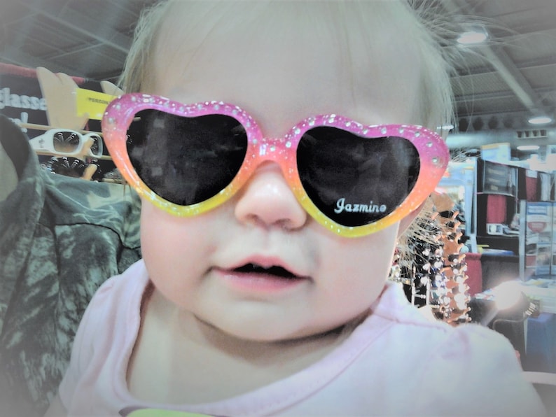 May include: A young girl wearing pink and yellow heart-shaped sunglasses with the word "Jazmino" printed on the lenses.