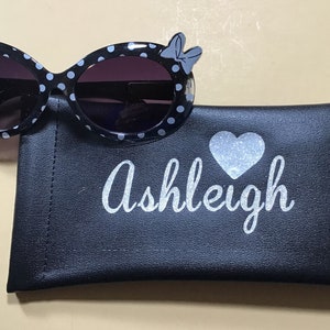 May include: Black and white polka dot sunglasses with a blue bow on each side. The sunglasses come with a black carrying case that has a silver glitter heart and the name "Ashleigh" written on it.