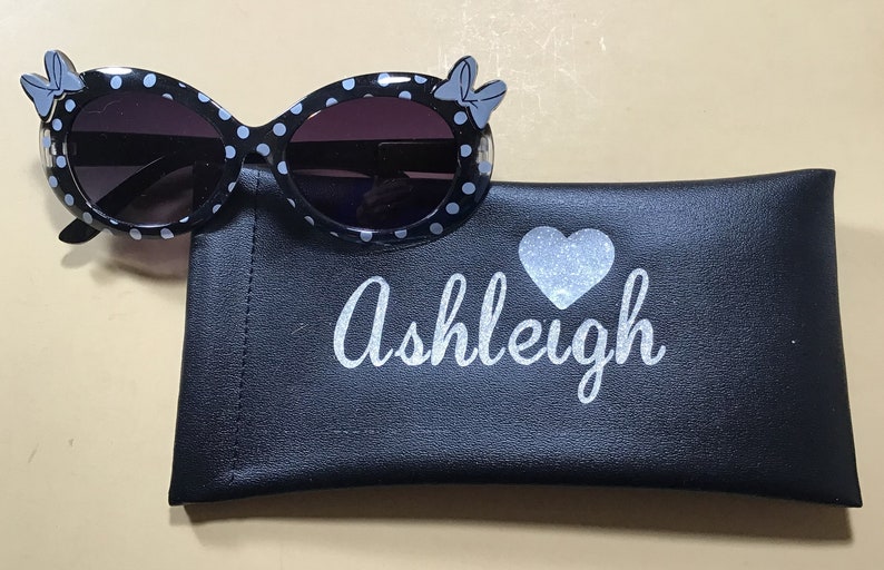 May include: Black and white polka dot sunglasses with a blue bow on each side. The sunglasses come with a black carrying case that has a silver glitter heart and the name "Ashleigh" written on it.