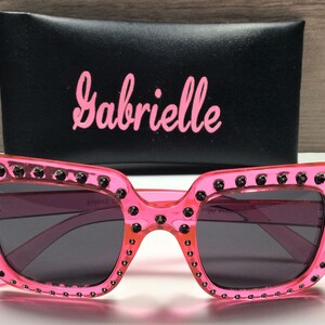 May include: Pink plastic sunglasses with black rhinestones around the frame and black lenses. The sunglasses come with a black case that has the word "Gabrielle" printed on it in pink.