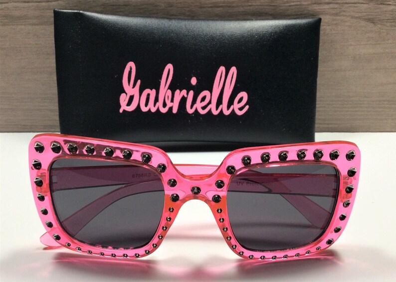 May include: Pink plastic sunglasses with black rhinestones around the frame and black lenses. The sunglasses come with a black case that has the word "Gabrielle" printed on it in pink.