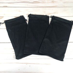 May include: Three black drawstring pouches made of soft fabric. The pouches are lying flat on a white wooden surface.