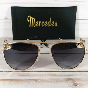 Op de afbeelding: Gouden aviator zonnebril met zwarte glazen en een vlinderontwerp aan elke kant. De zonnebril ligt op een wit houten oppervlak. De zonnebril wordt geleverd met een zwarte etui met daarop "Mercedes".