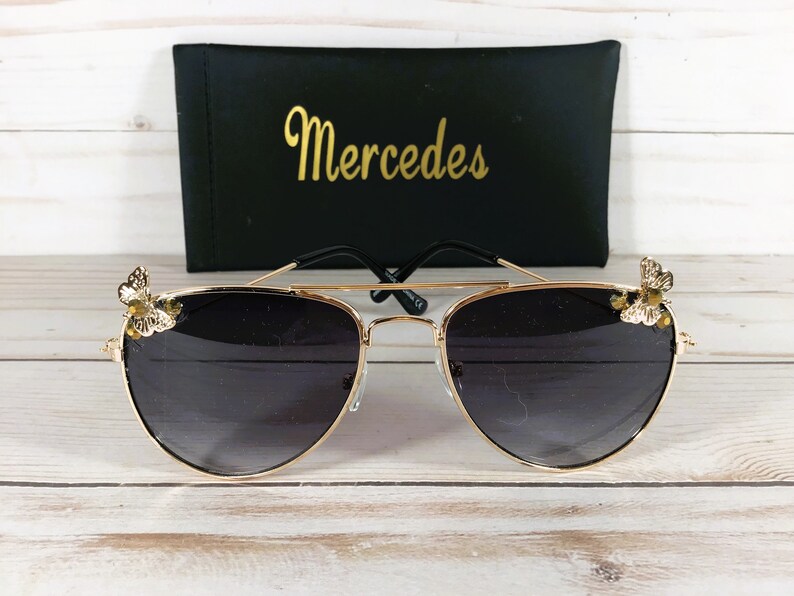 Op de afbeelding: Gouden aviator zonnebril met zwarte glazen en een vlinderontwerp aan elke kant. De zonnebril ligt op een wit houten oppervlak. De zonnebril wordt geleverd met een zwarte etui met daarop "Mercedes".