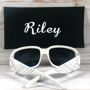 May include: White sunglasses with dark lenses and decorative accents on the temples. The sunglasses are paired with a black case that has the name "Riley" in white script.