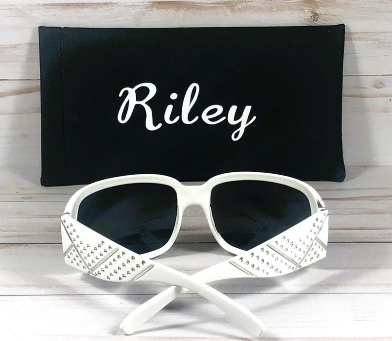 May include: White sunglasses with dark lenses and decorative accents on the temples. The sunglasses are paired with a black case that has the name "Riley" in white script.
