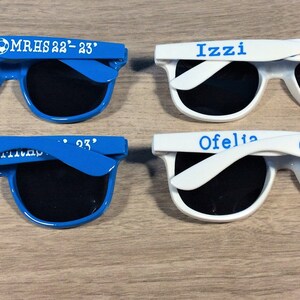 May include: Four pairs of sunglasses with personalized text and a soccer ball graphic. Two pairs are blue and two pairs are white.