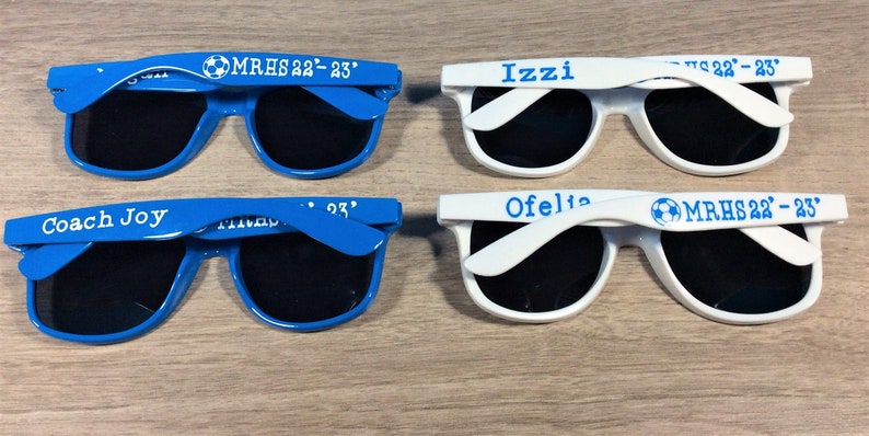 May include: Four pairs of sunglasses with personalized text and a soccer ball graphic. Two pairs are blue and two pairs are white.