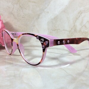 May include: Cat-eye style eyeglasses with a pink and tortoiseshell pattern frame. The glasses have clear lenses and are embellished with rhinestones on the frame. The temples are a dark brown color. A pink flower is in the background.