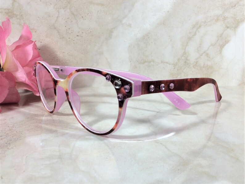 May include: Cat-eye style eyeglasses with a pink and tortoiseshell pattern frame. The glasses have clear lenses and are embellished with rhinestones on the frame. The temples are a dark brown color. A pink flower is in the background.