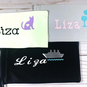 May include: Three different eyeglass cases with the name "Liza" embroidered on them. The first case is white with a purple cat with a heart on it. The second case is gray with the name "Liza" in pink letters and two blue wine glasses. The third case is black with a white ship and the name "Liza" in white cursive.