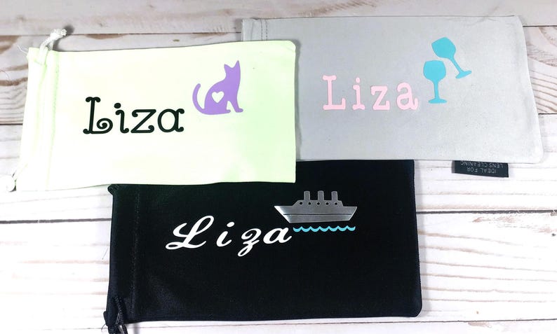 May include: Three different eyeglass cases with the name "Liza" embroidered on them. The first case is white with a purple cat with a heart on it. The second case is gray with the name "Liza" in pink letters and two blue wine glasses. The third case is black with a white ship and the name "Liza" in white cursive.
