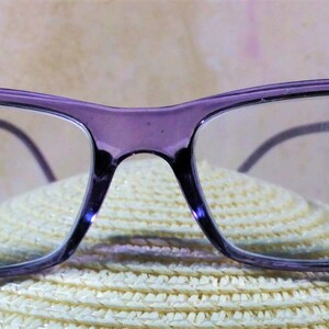 May include: Rectangular eyeglasses with a translucent purple frame. The outer corners of the frame feature small, dark purple decorative rhinestones. The lenses are clear and the temples are thin, matching the purple color of the front frame.