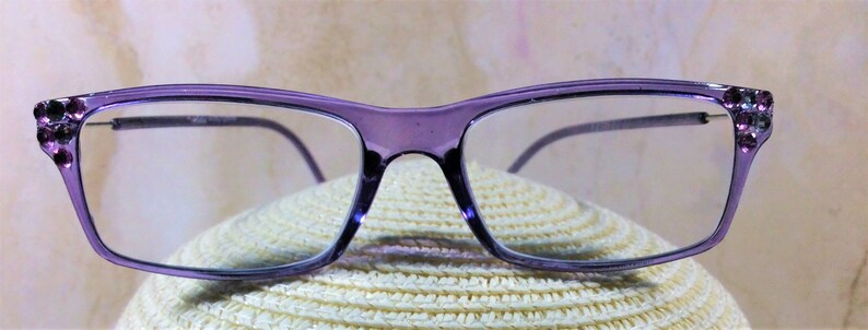 May include: Rectangular eyeglasses with a translucent purple frame. The outer corners of the frame feature small, dark purple decorative rhinestones. The lenses are clear and the temples are thin, matching the purple color of the front frame.