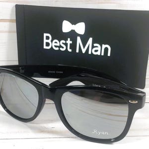 May include: Black sunglasses with mirrored lenses and a black case. The case has a white bow tie and the text "Best Man". The sunglasses have the name "Ryan" printed on the right lens.