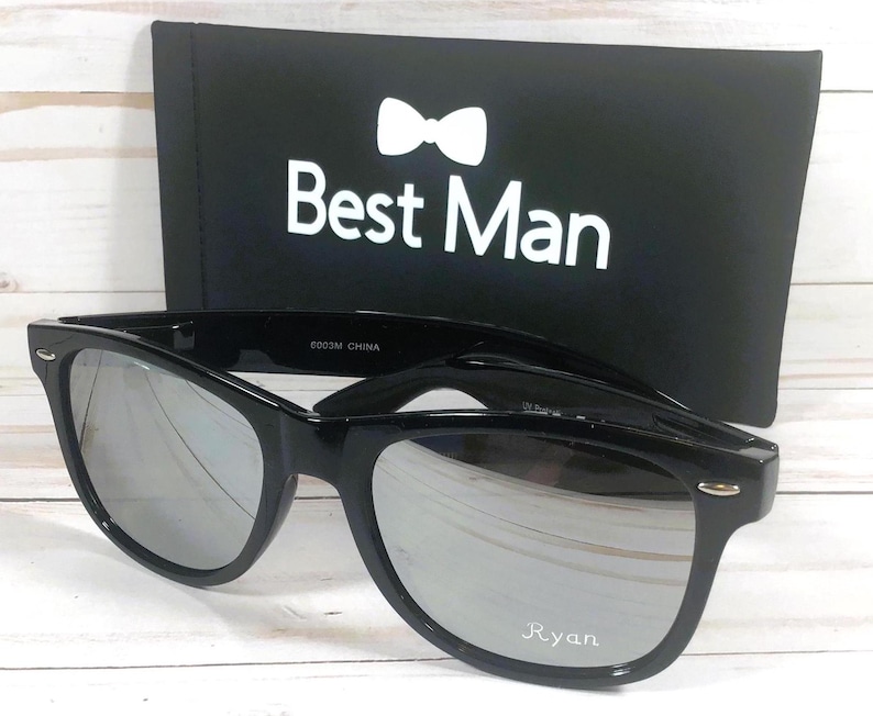 May include: Black sunglasses with mirrored lenses and a black case. The case has a white bow tie and the text "Best Man". The sunglasses have the name "Ryan" printed on the right lens.