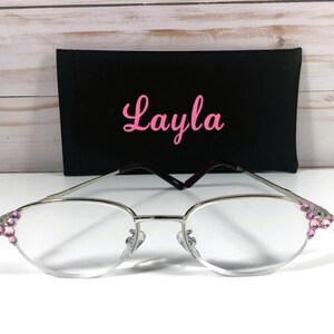May include: Silver-framed eyeglasses with pink jewel accents on the corners, resting in front of a black case. The case has the name "Layla" in pink cursive script. The glasses have a classic oval shape.