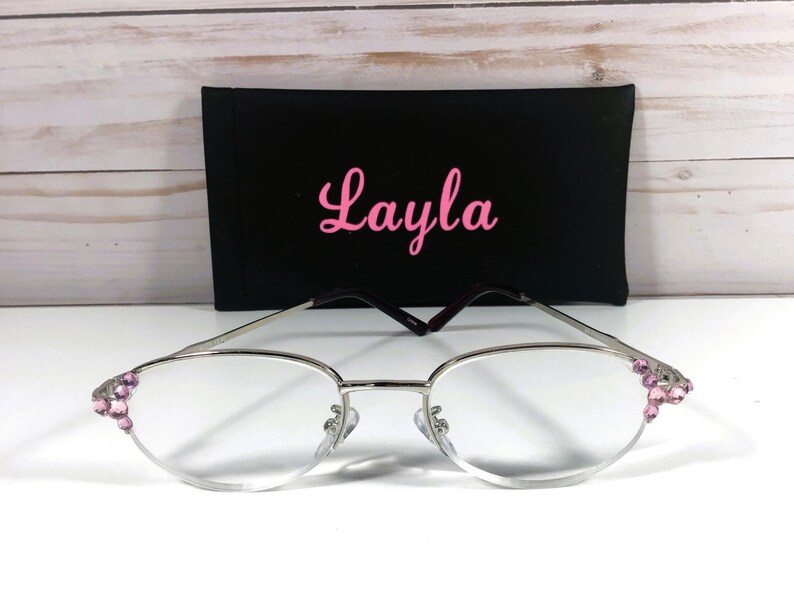 May include: Silver-framed eyeglasses with pink jewel accents on the corners, resting in front of a black case. The case has the name "Layla" in pink cursive script. The glasses have a classic oval shape.