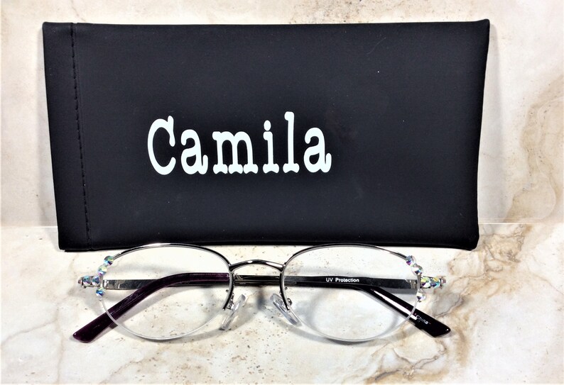 May include: Silver-framed spectacles with a black case. The spectacles have a half-rim design with clear lenses and purple arms. The case is black with the name "Camila" in white lettering. The spectacles have decorative rhinestones.