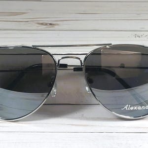 May include: Silver-framed aviator sunglasses with gradient lenses. The lenses transition from dark to light gray. The name "Alexander" is printed on the right lens. The sunglasses are set against a white wooden background.