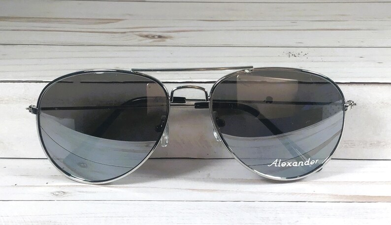 May include: Silver-framed aviator sunglasses with gradient lenses. The lenses transition from dark to light gray. The name "Alexander" is printed on the right lens. The sunglasses are set against a white wooden background.