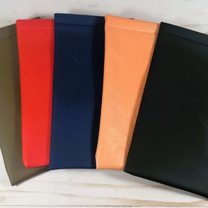 May include: A collection of five colorful eyeglass cases in various shades. The cases are arranged side by side, showcasing colors like olive green, red, navy blue, orange, and black. The cases are made of a smooth material.