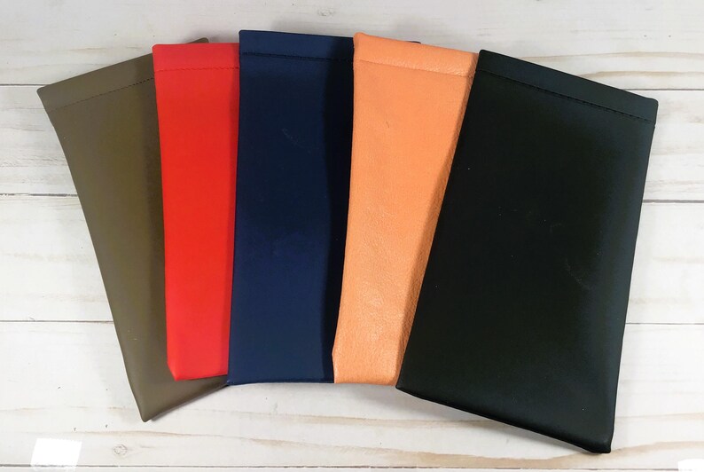 May include: A collection of five colorful eyeglass cases in various shades. The cases are arranged side by side, showcasing colors like olive green, red, navy blue, orange, and black. The cases are made of a smooth material.