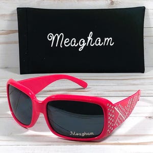 K&ouml;nnte beinhalten: Rosa Sonnenbrille mit schwarzen Gl&auml;sern und Strasssteinen an den B&uuml;geln. Die Sonnenbrille hat den Schriftzug "Meagham" auf den Gl&auml;sern. Die Sonnenbrille befindet sich in einem schwarzen Etui mit dem Schriftzug "Meagham" auf der Vorderseite.