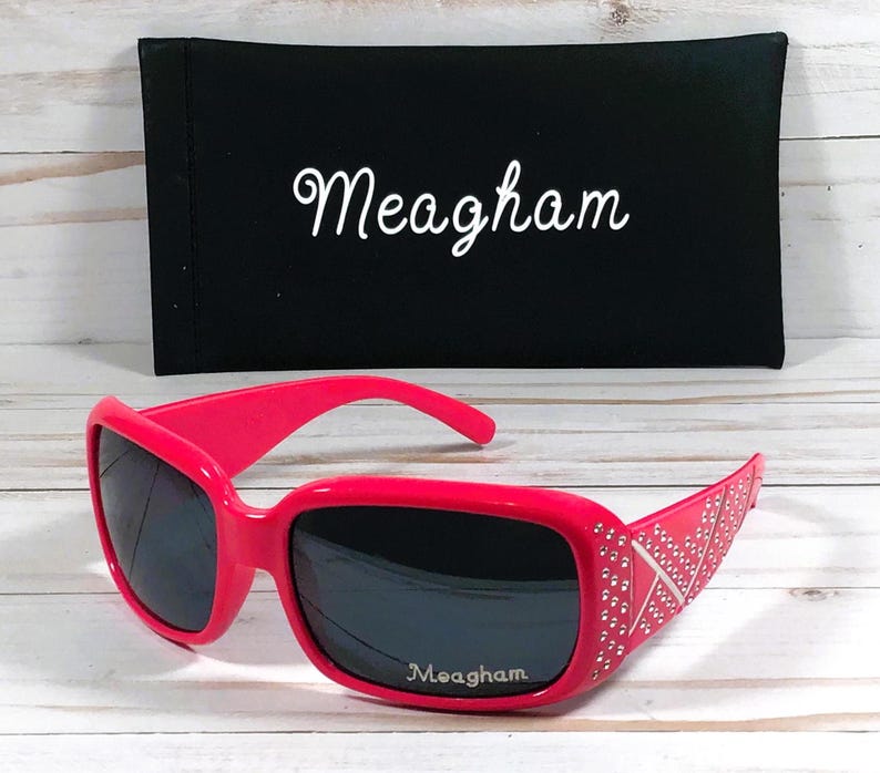 K&ouml;nnte beinhalten: Rosa Sonnenbrille mit schwarzen Gl&auml;sern und Strasssteinen an den B&uuml;geln. Die Sonnenbrille hat den Schriftzug "Meagham" auf den Gl&auml;sern. Die Sonnenbrille befindet sich in einem schwarzen Etui mit dem Schriftzug "Meagham" auf der Vorderseite.