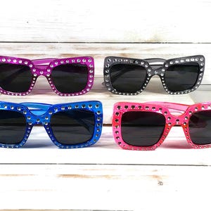 May include: Four pairs of sunglasses with rhinestones around the frames. The sunglasses are in four different colors: pink, blue, black, and purple.